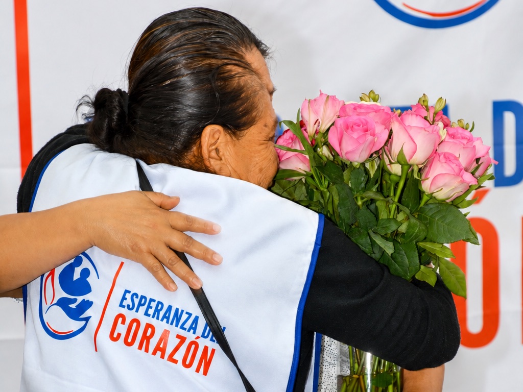 Una adulta mayor con un ramo de rosas recibe un abrazo durante un reencuentro familiar