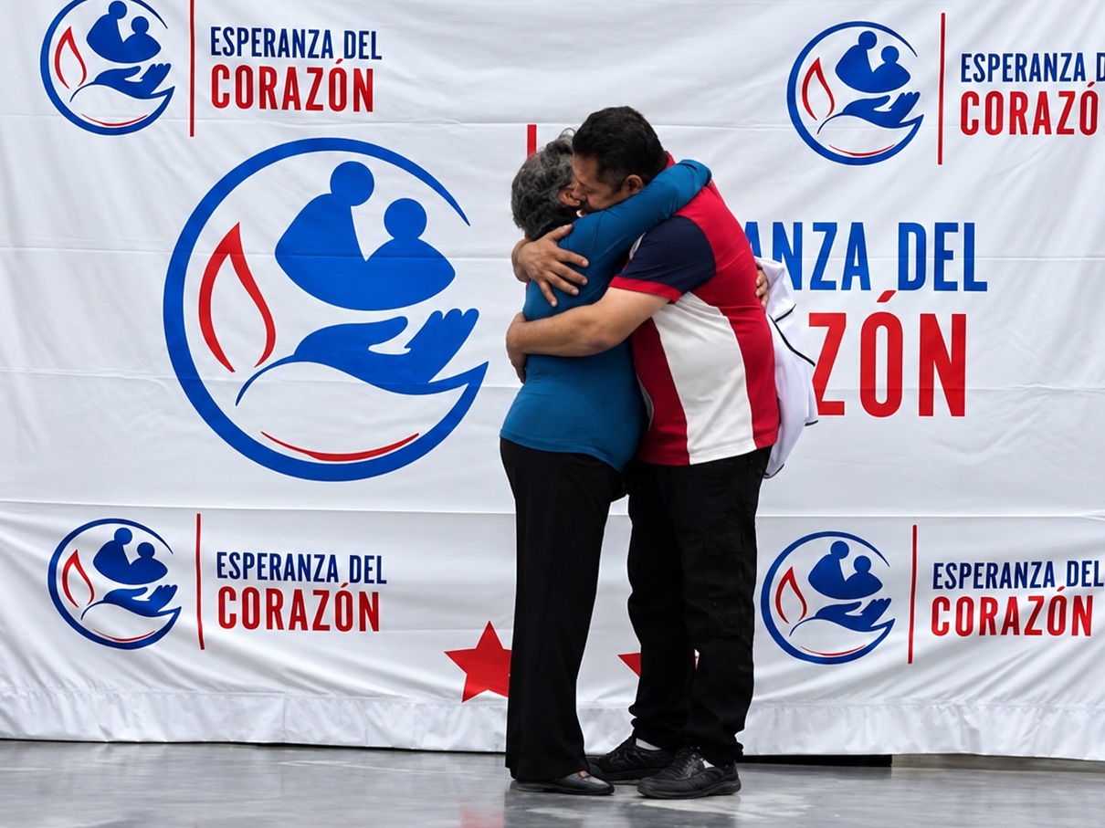 Dos personas se abrazan frente al cartel de Esperanza del Corazón durante un reencuentro familiar